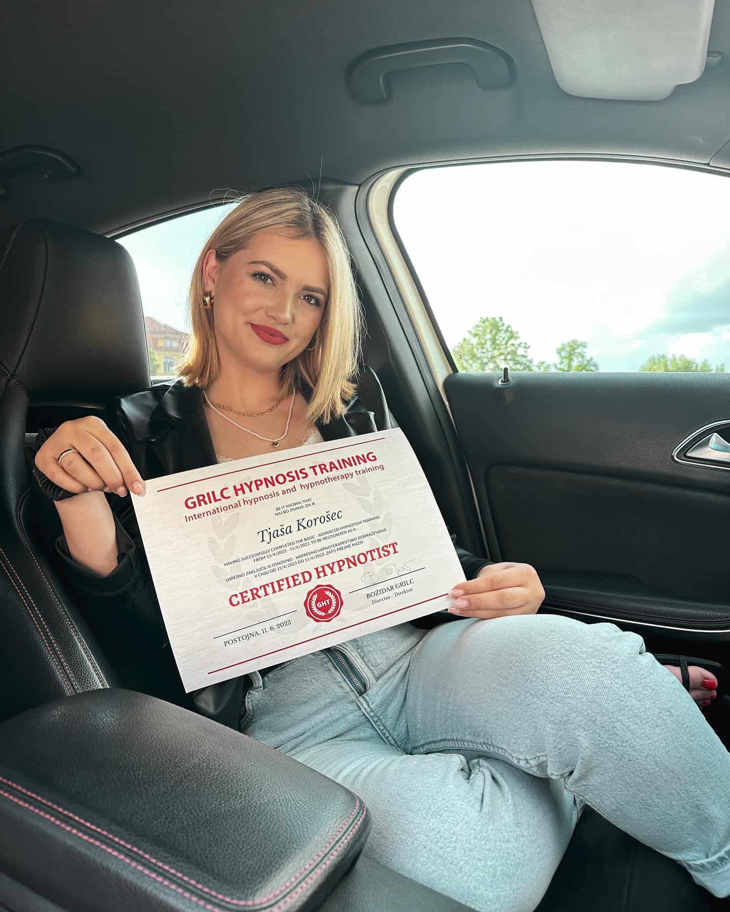 Certified hypnotist holding diploma for hypnotherapy training in a car, showcasing expertise in hypnotherapy, hypnosis, and personal development. Woman with blonde hair, red lipstick, and casual attire.